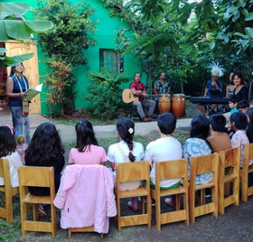 Kinder sitzen draußen auf Stühlen und hören einer Erzählerin und einer Musikgruppe zu